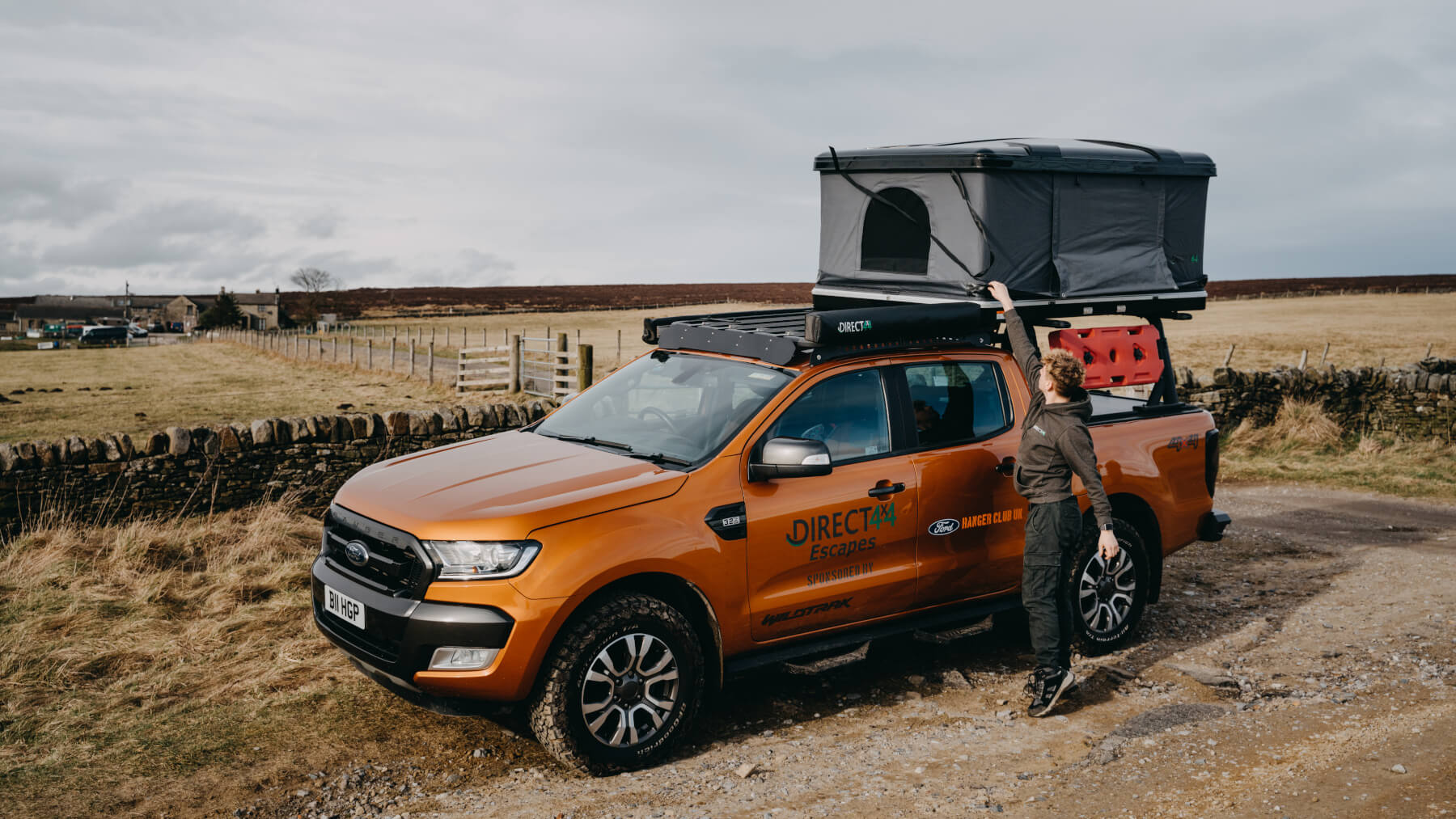 Photo of an orange Ford Ranger Wildtrak pickup truck in a countryside farmer's field equipped with Direct4x4 overland expedition gear including roof tack, rooftop camping tent and more.