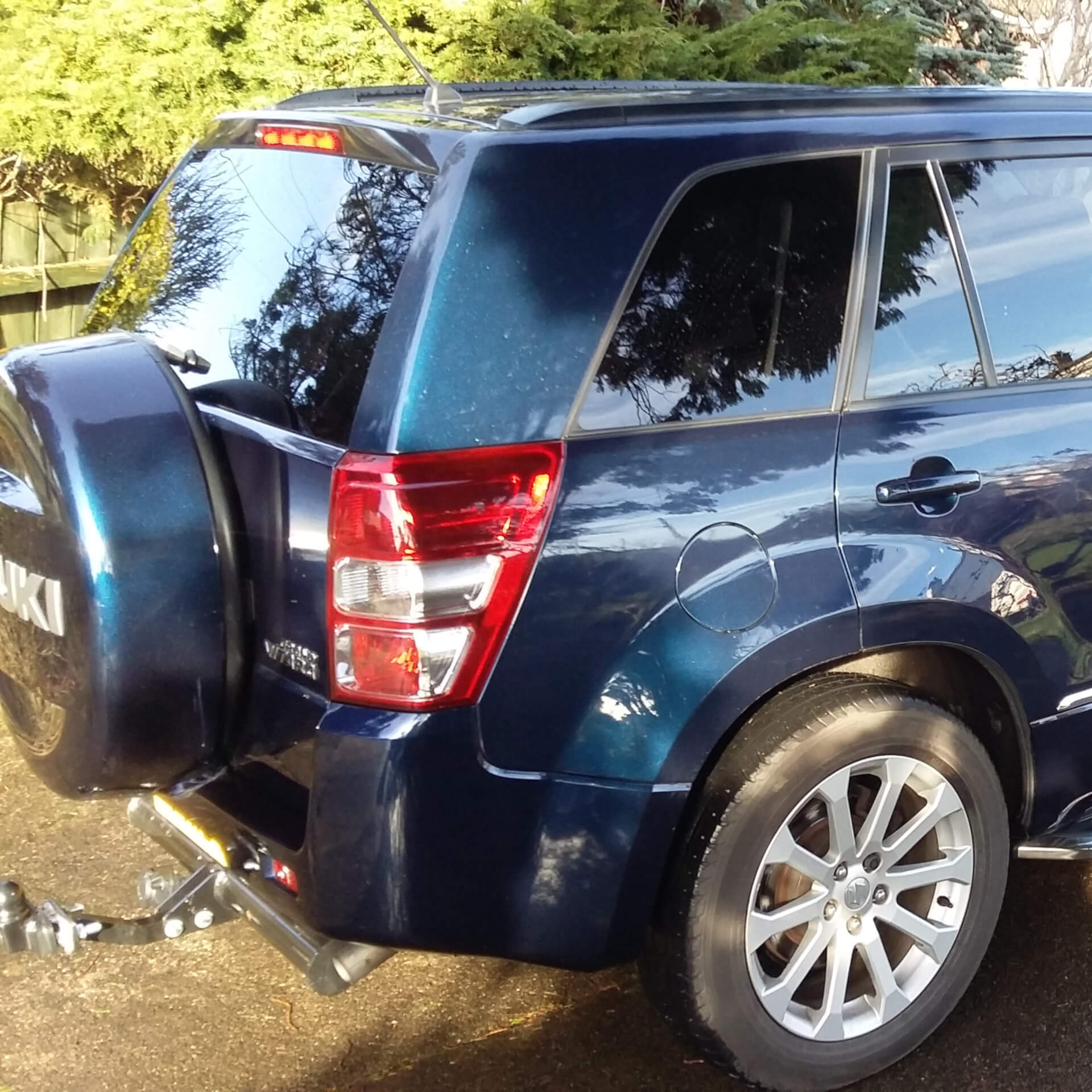 Direct4x4 accessories for Suzuki Grand Vitara vehicles with a photo of the back of a blue Suzuki Grand Vitara with the spare tyre on the back parked in front of trees