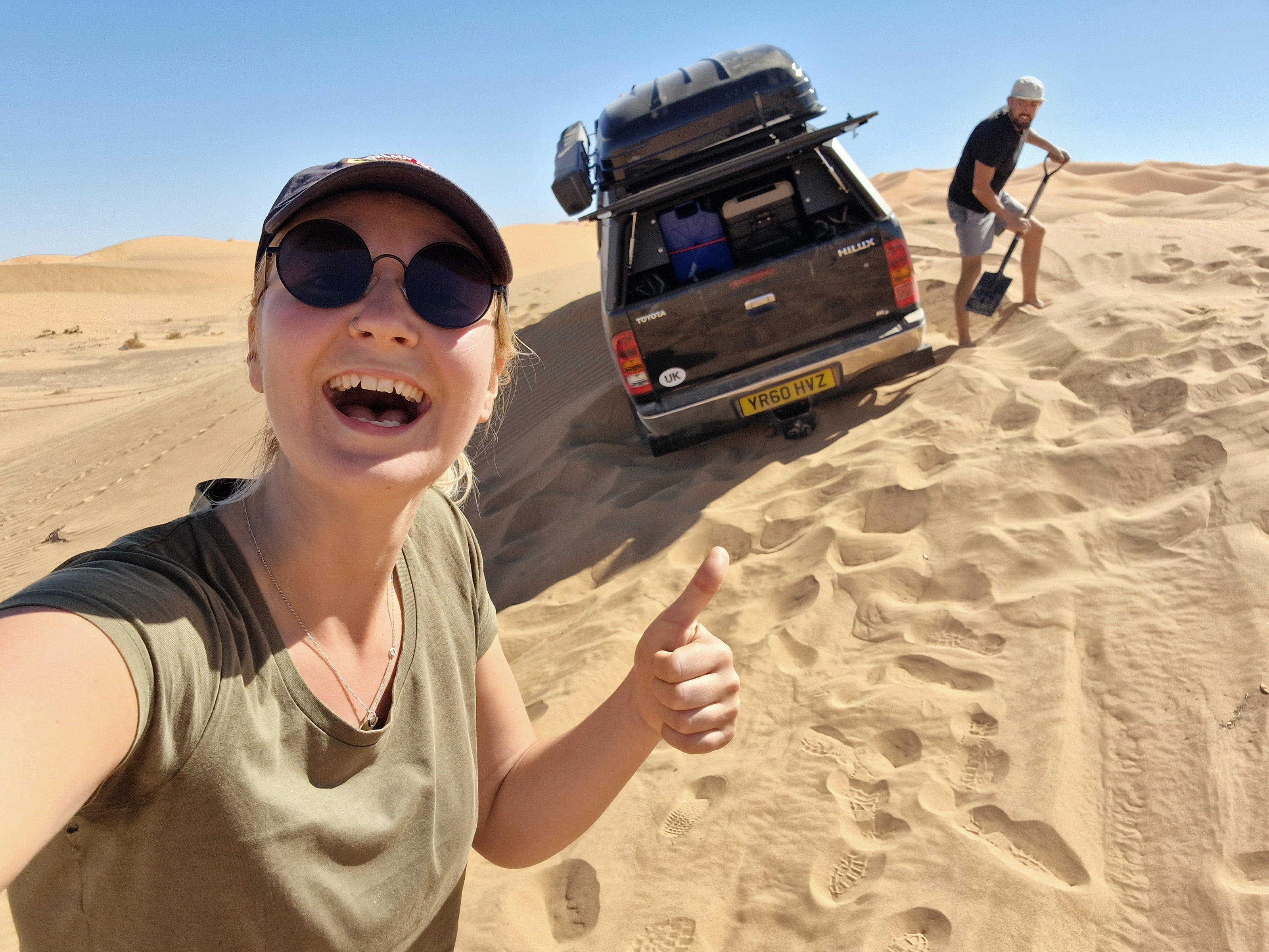 Photo of 2 people trying to dig out a Toyota Hilux pickup truck from the Moroccan desert.