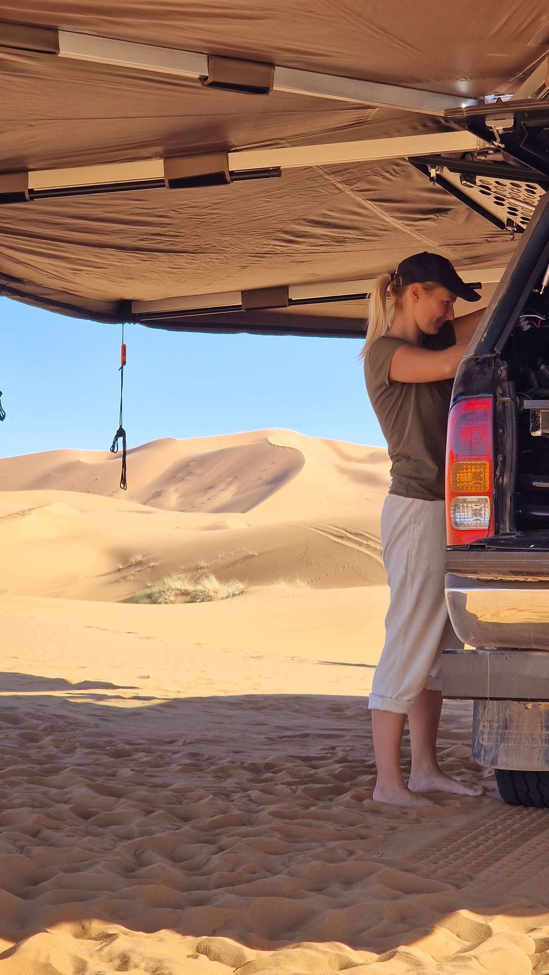 Photo of woman cooking out the back of a pickup truck load bed tailgate under a vehicle side awning in the evening in the Moroccan desert.