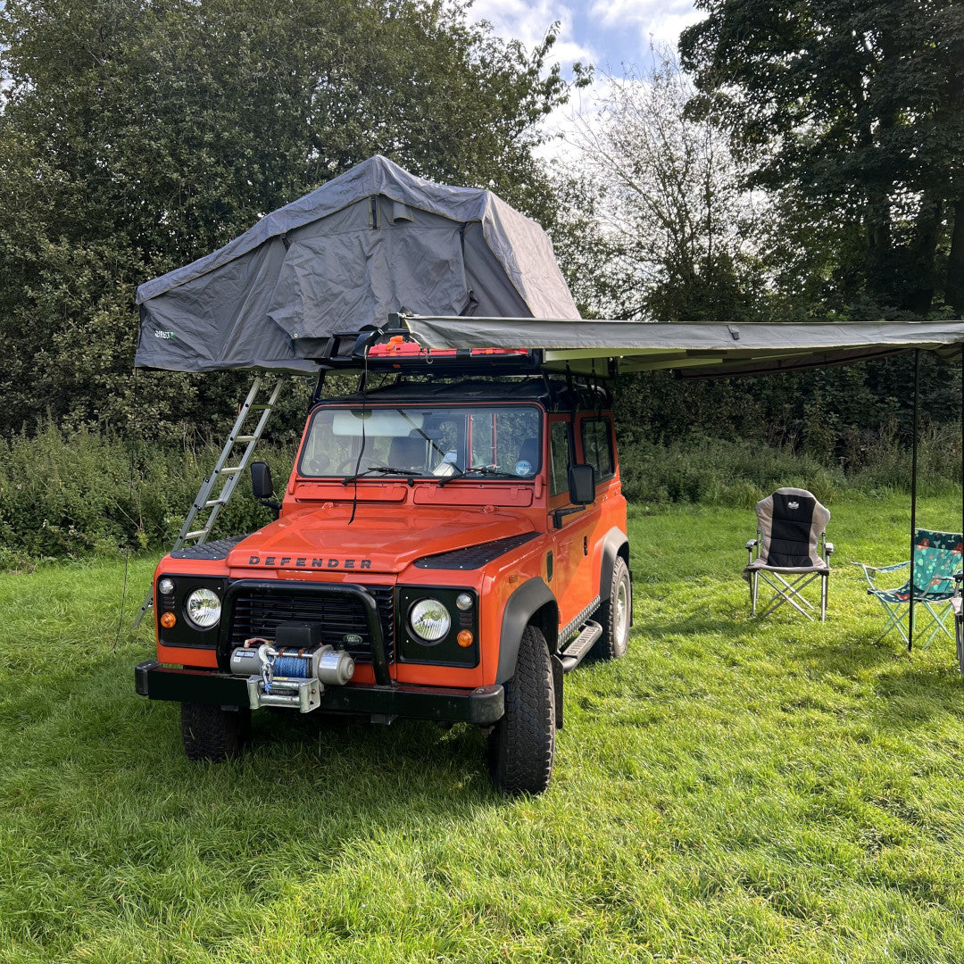 Photo off an orange Land Rover Defender 90 with a Direct4x4 roof top expedition camping tent and overland vehicle side awning in a field.