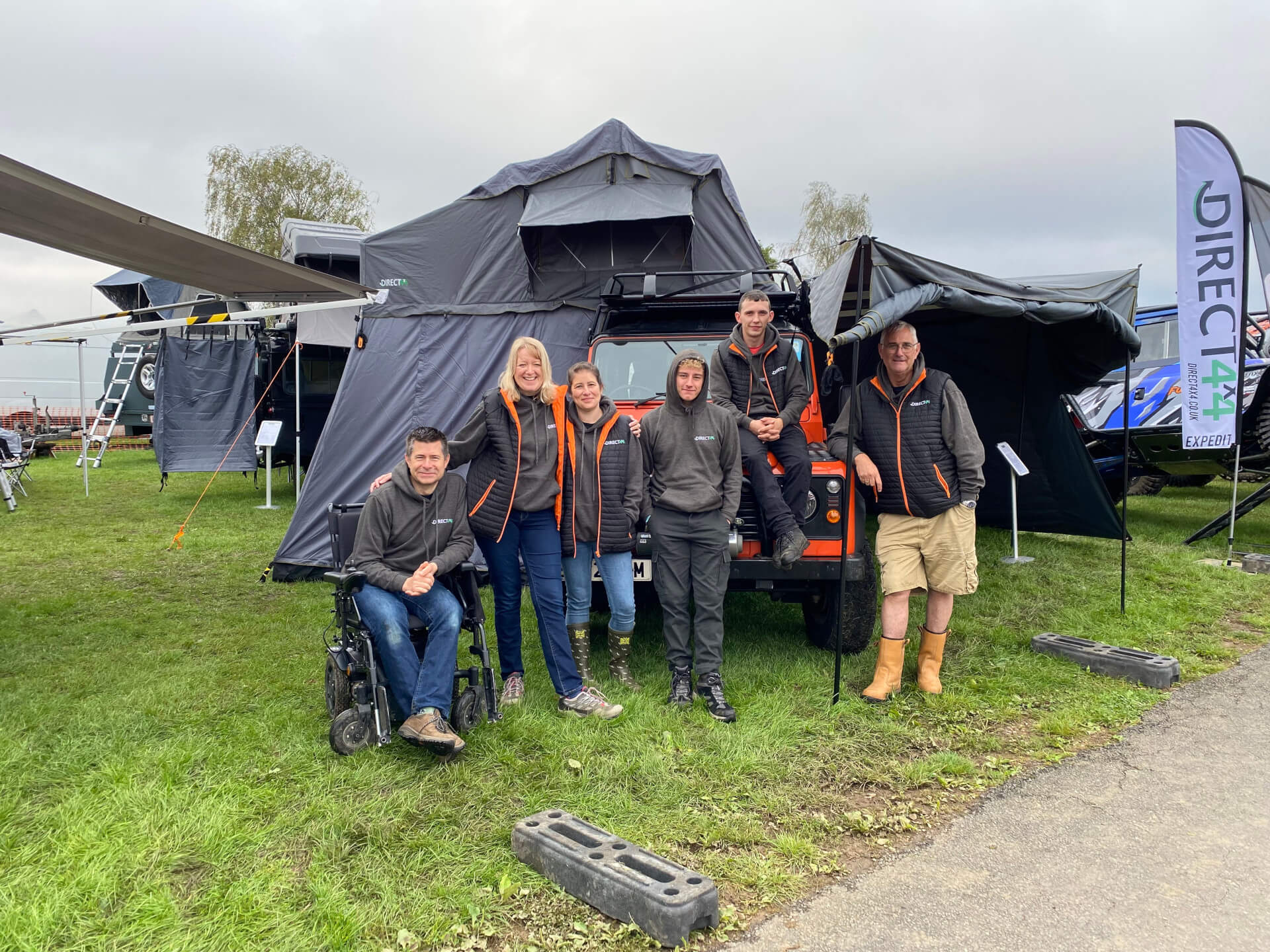 Photo of the Direct4x4 team at a showground in front of an organe Land Rover Defender with a camping tent.
